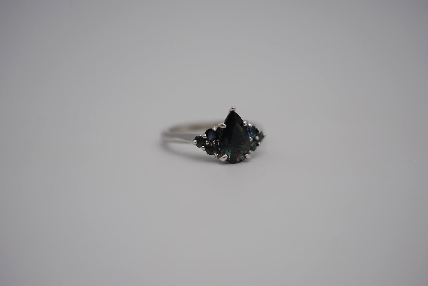 A white gold ring with a pear cut blue green sapphire with three round sapphire sidestones to each side in a triangle. 